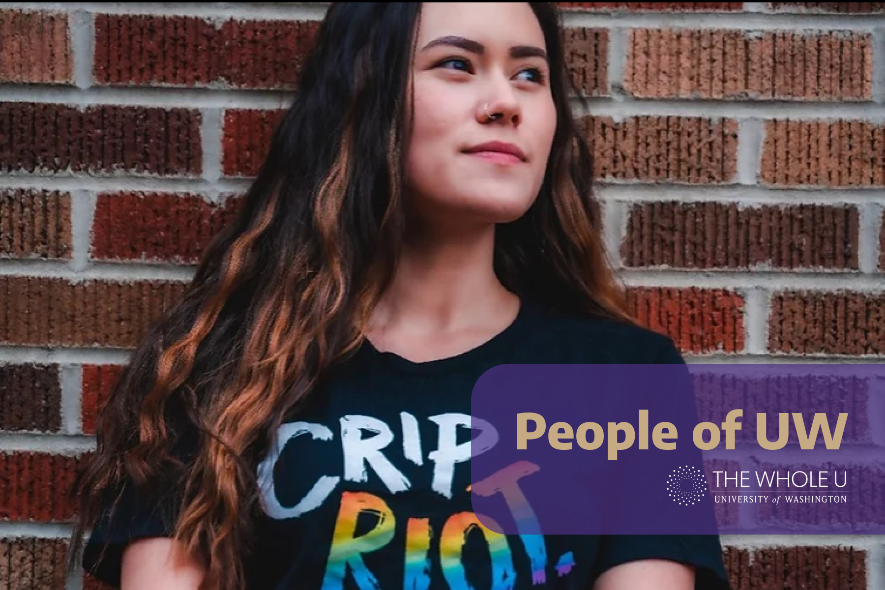 Asian American woman with long wavy brown hair, wearing a black shirt with rainbow text that reads "CRIP RIOT", looks away from the camera, against a brick wall.
