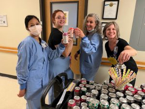 Four healthcare workers, two in scrubs and two in Project Recharge 2.0 t-shirts, are gathered around a refreshment cart filled with beverages. They are holding up cans and smiling as they enjoy the refreshments.