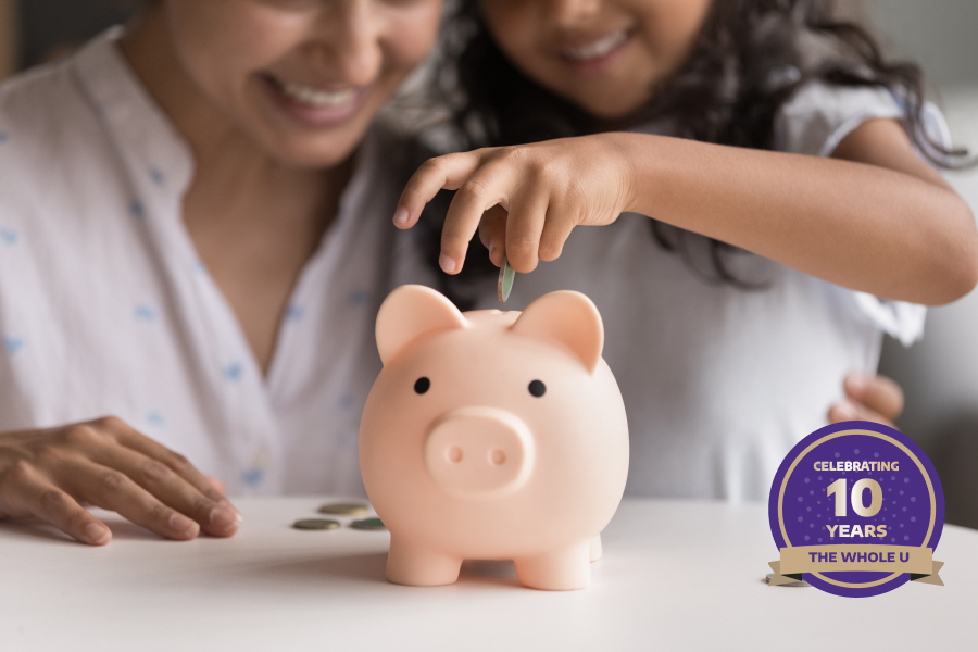 A child and an adult smiling as the child places a coin into a pink piggy bank, symbolizing teaching kids about saving money.