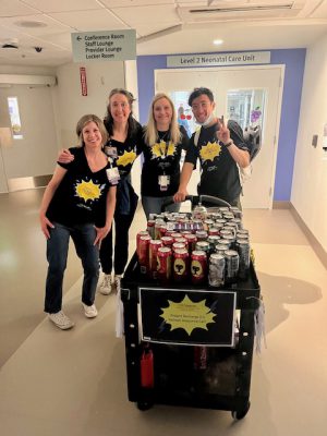 A group of four staff members, wearing black t-shirts with the Project Recharge 2.0 logo, stand around a refreshment cart. They are in a hallway near the entrance to a Neonatal Care Unit, smiling and posing for the camera. The cart is filled with assorted beverages.