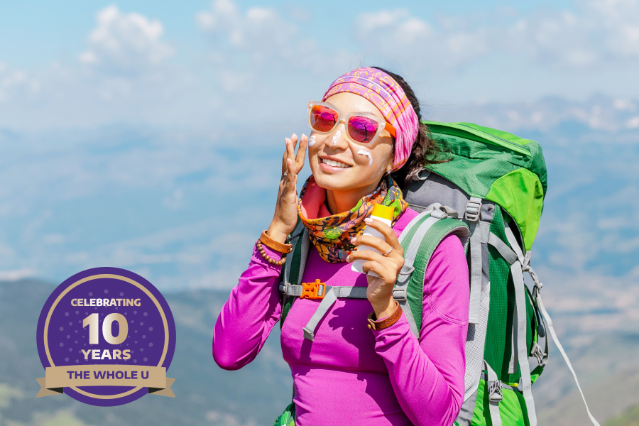 A women hiker in a pink top and green backpack applies sunscreen to her face.
